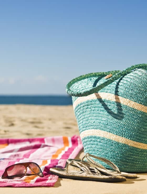 A beach scene featuring a woven tote, sunglasses, flip-flops, and a striped towel on the sand.