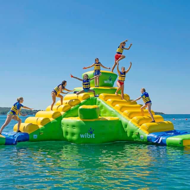 A group of children in life vests play and climb on a colorful inflatable water attraction in bright blue waters.