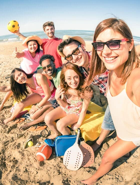 A group of eight friends joyfully posing for a selfie on the beach with sports equipment and a clear sky in the background.