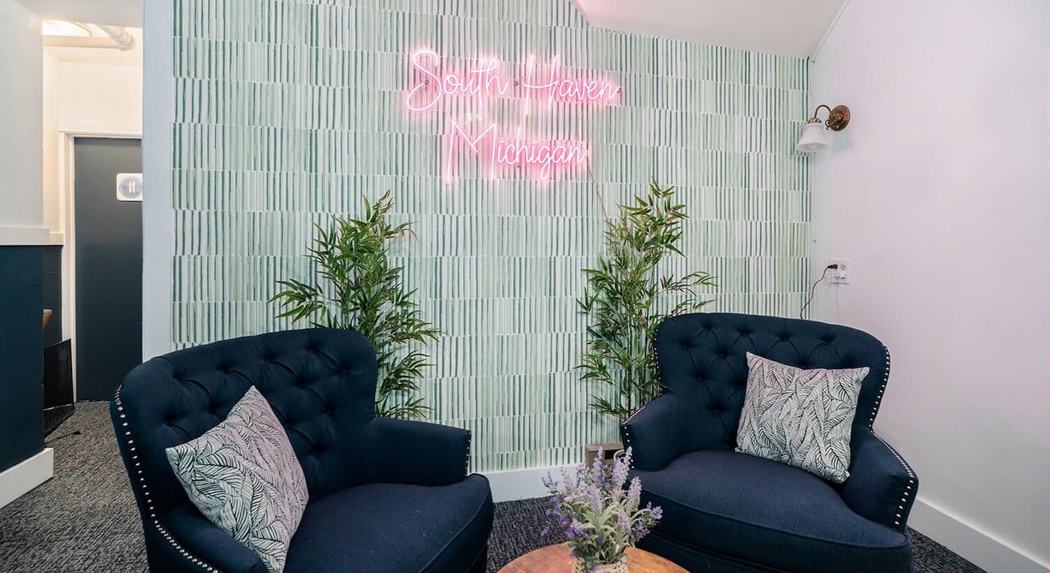 A cozy seating area featuring two dark blue armchairs, a wooden table, and a neon sign on a patterned wall.